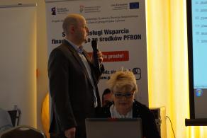 Powiększ obraz: dwie osoby na zdjeciu, jedna przemawia, druga pracuje przed komputerem